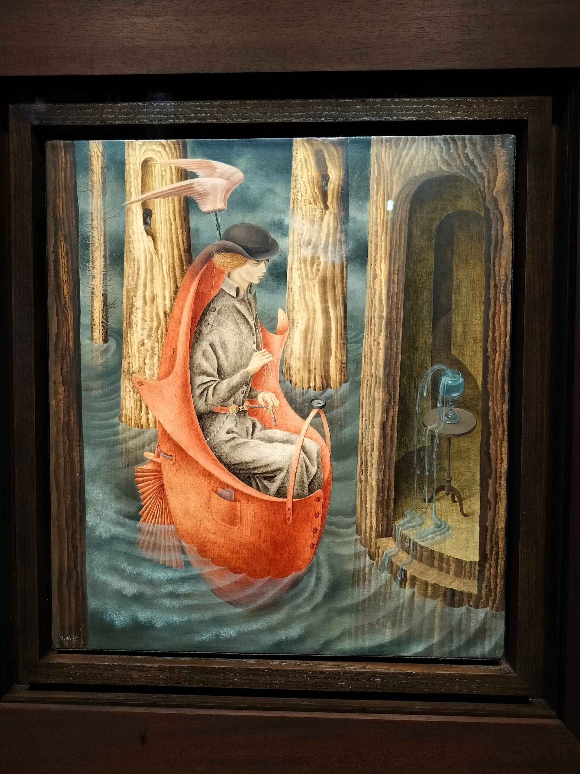 Remedios Varos maleri Exploración de las fuentes del río Orinoco: en kvinde i trenchcoat og bowlerhat sejler i en rød, ægformet båd syet af en frakke gennem en oversvømmet skov mod et hulrum i en træstamme, hvor et bæger løber over med vand.