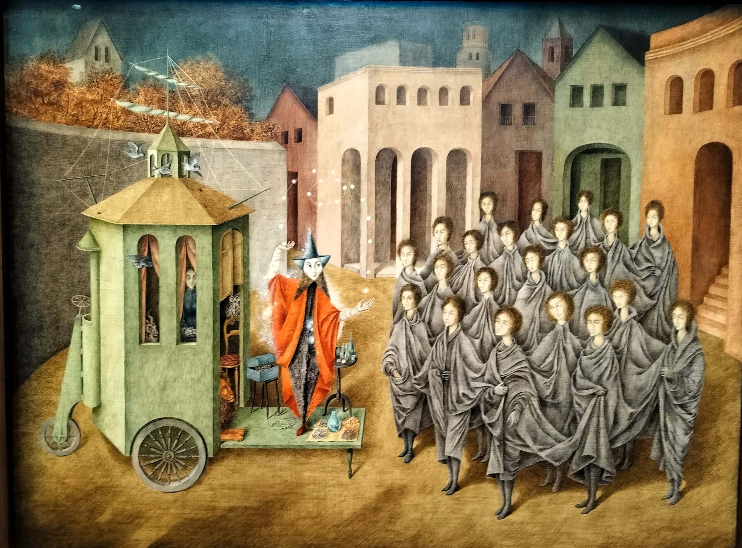 Remedios Varos maleri El juglar: en tryllekunstner i rød kappe og spids hat viser kunster frem foran en grøn vogn på hjul for en tæt flok ensartede figurer i grå kapper på en torveplads.